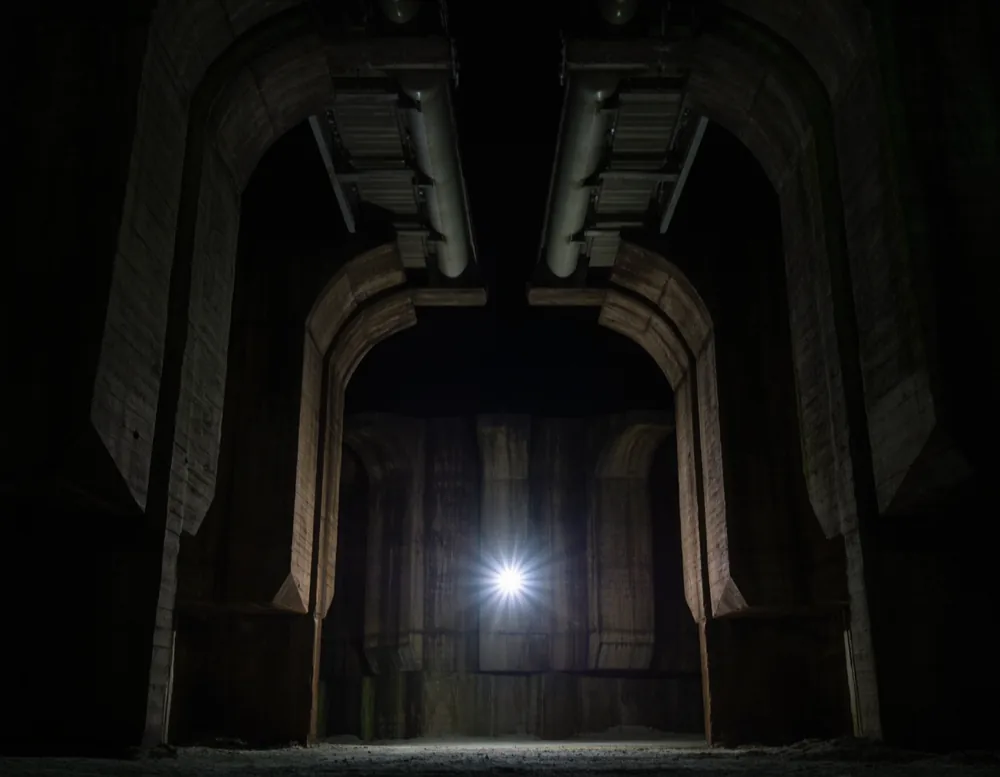 A vast raw concrete bunker interior with conveyor bridges overhead, a single shaft of light falling to the floor.