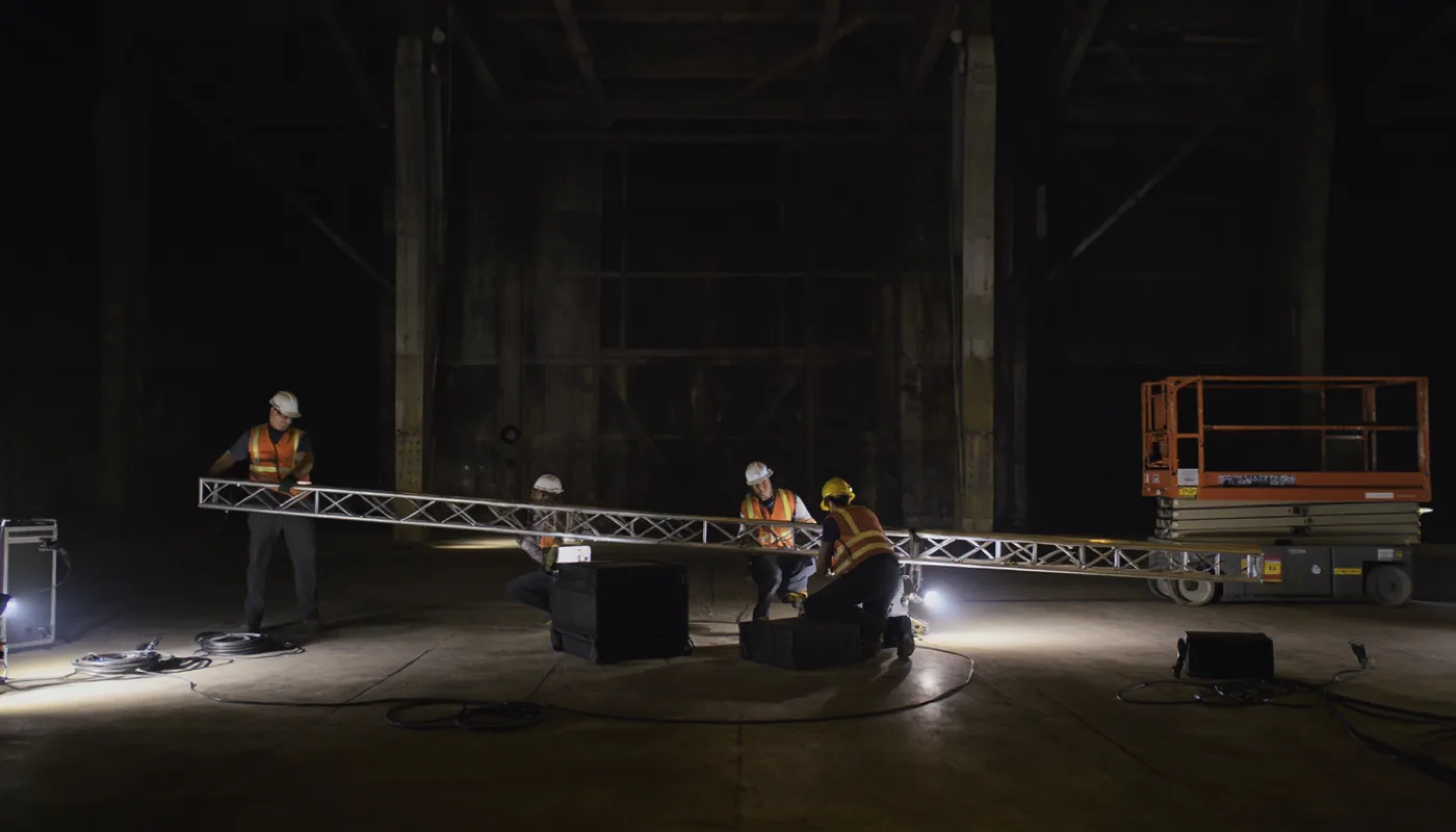 Event technicians in hard hats and hi-vis vests assembling an aluminium truss in a dark industrial hall.