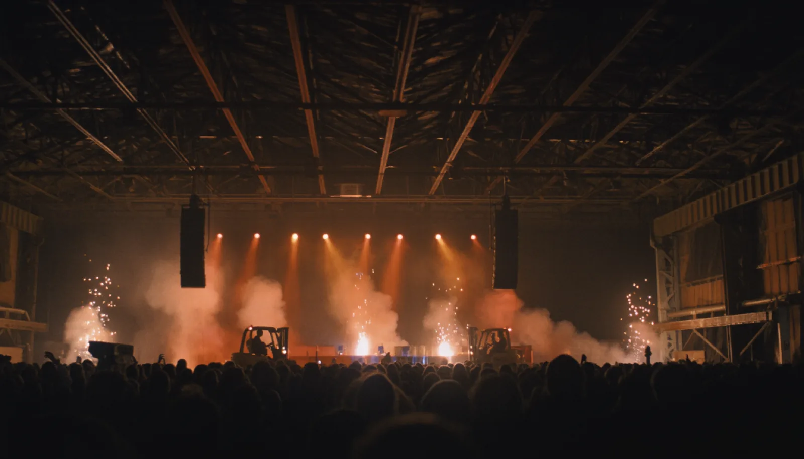 A dense crowd in a dark industrial hall in front of a low stage, sparks falling behind the performers.
